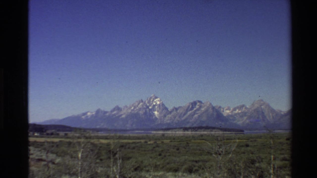 Wide 1980 home-movie frame of Rocky Mountain peaks seen across low grassy terrain and scattered bare trees.