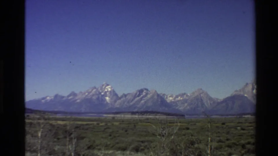 1980 8mm landscape frame of snow-capped Western U.S. peaks over a flat green-brown lowland.