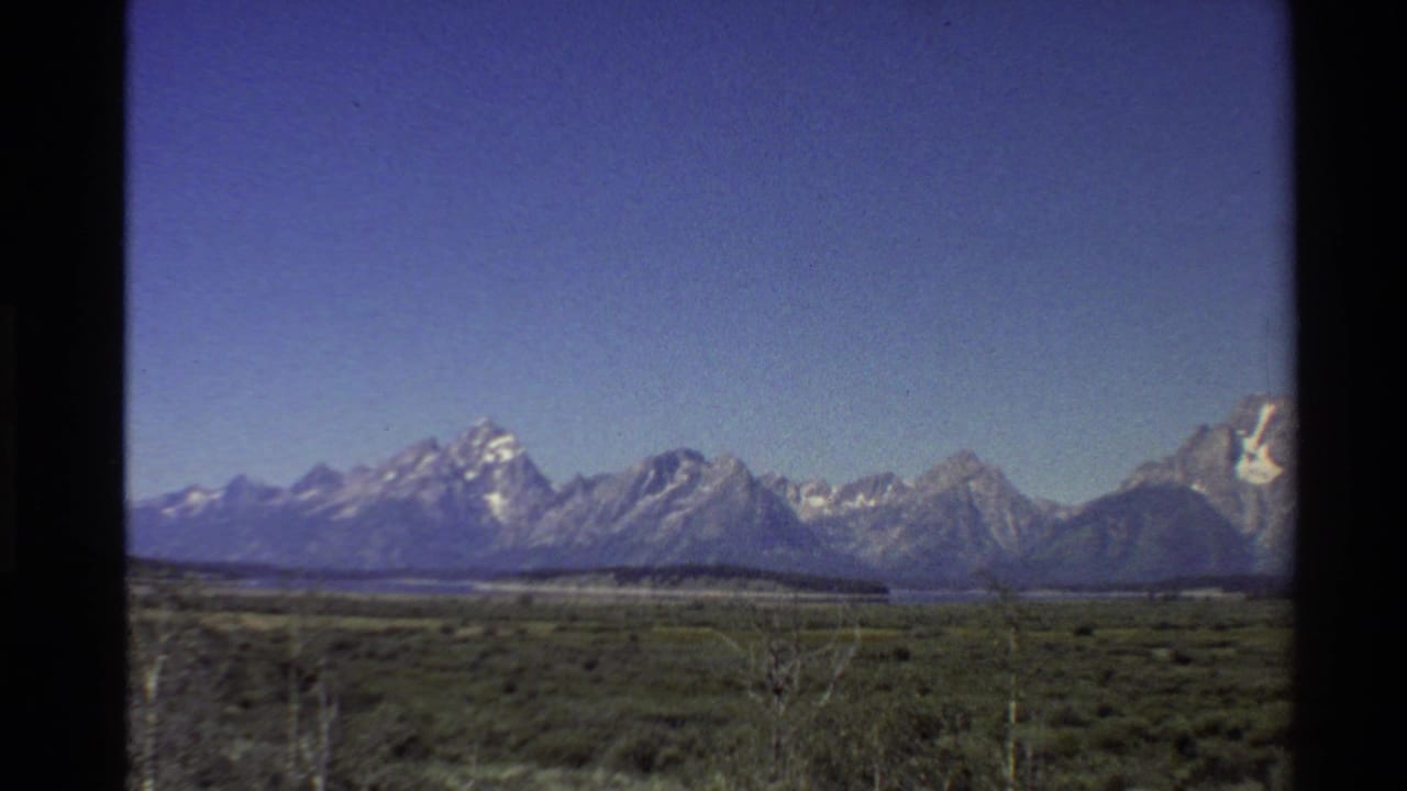 1980 8mm Western USA home-movie frame showing snowy mountains across open plain vegetation under a clear blue sky.