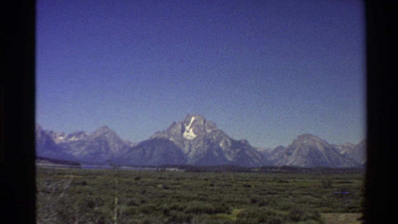 1980 8mm view of the Teton Range rising beyond low green valley vegetation under a clear blue sky.
