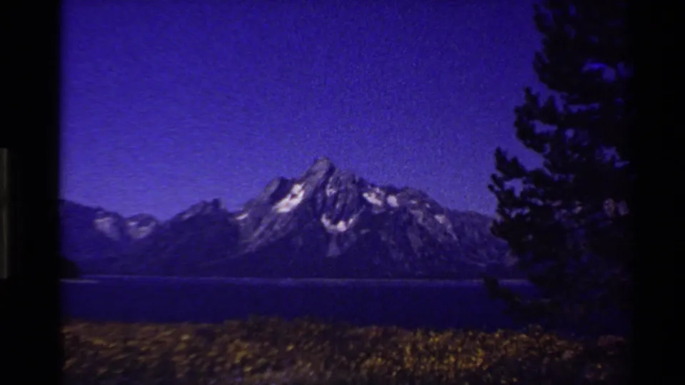 1980 Western USA 8mm view of snow-streaked mountain peaks above a lake, with wildflowers below and a dark pine at right.