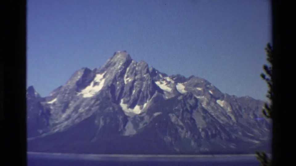 Closer 8mm zoom view of a rocky Western U.S. mountain face with lingering snowfields above a blue lake.