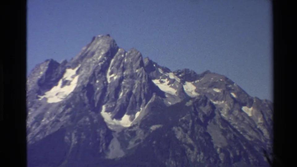 Tight 1980 8mm view of a jagged Western U.S. mountain peak with snow patches, rocky slopes, and pine branches at the right edge.