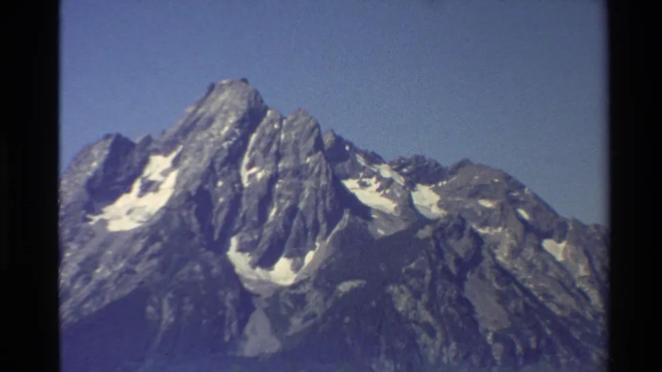 Soft 1980 8mm travel frame showing snow-marked rocky mountain peaks against a clear blue sky in the Western United States.