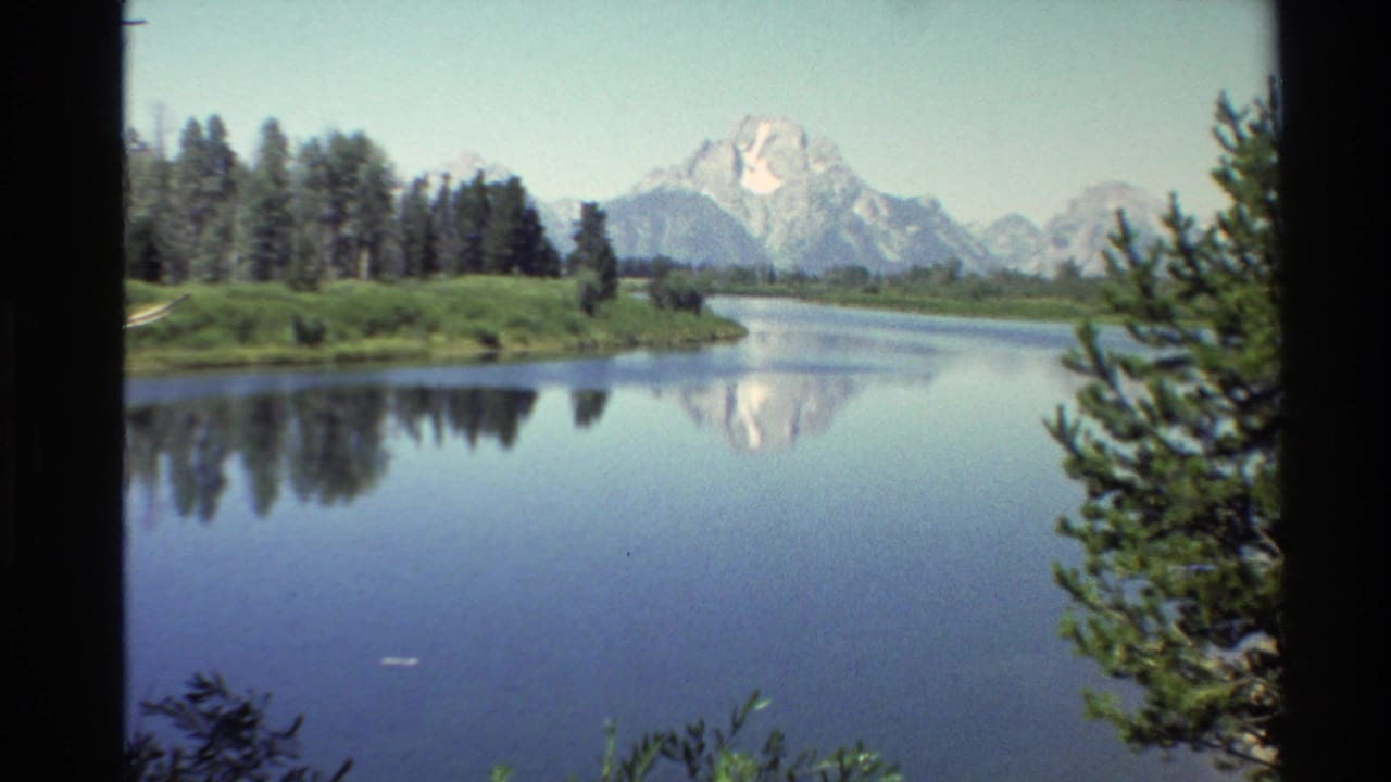 1980 8mm Western USA landscape view of a still river reflecting snow-capped mountains, with conifers and grassy banks under a clear sky.