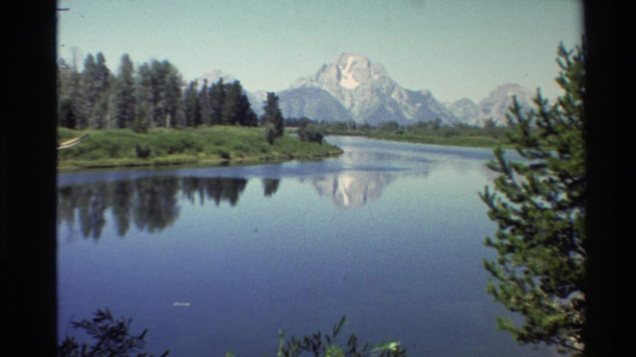 Western USA 8mm frame from 1980 showing a river bend, conifer-lined shore, distant snowy mountains, and their soft reflection in the water.