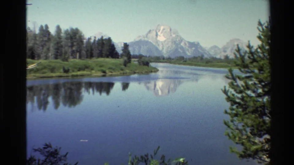 Western USA 8mm frame from 1980 showing a river bend, conifer-lined shore, distant snowy mountains, and their soft reflection in the water.