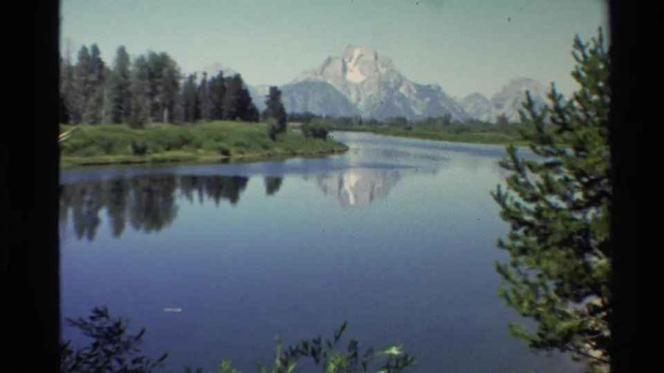 Western USA 1980 8mm landscape with a calm river, forested bank, snow-capped mountains, and pine branches partly framing the right side.
