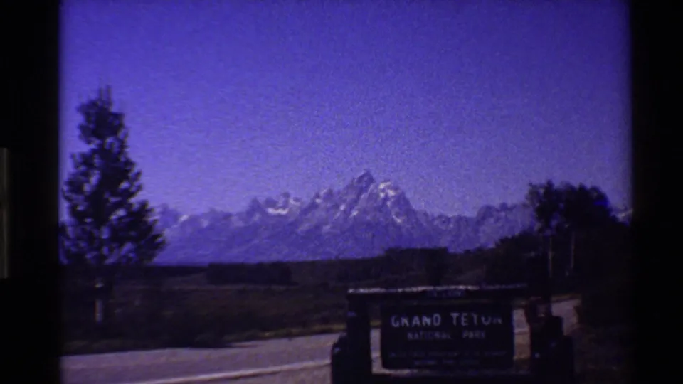 1980 8mm frame appears to show the Grand Teton National Park entrance sign beside a road, with the Teton range in the background and a strong purple cast.