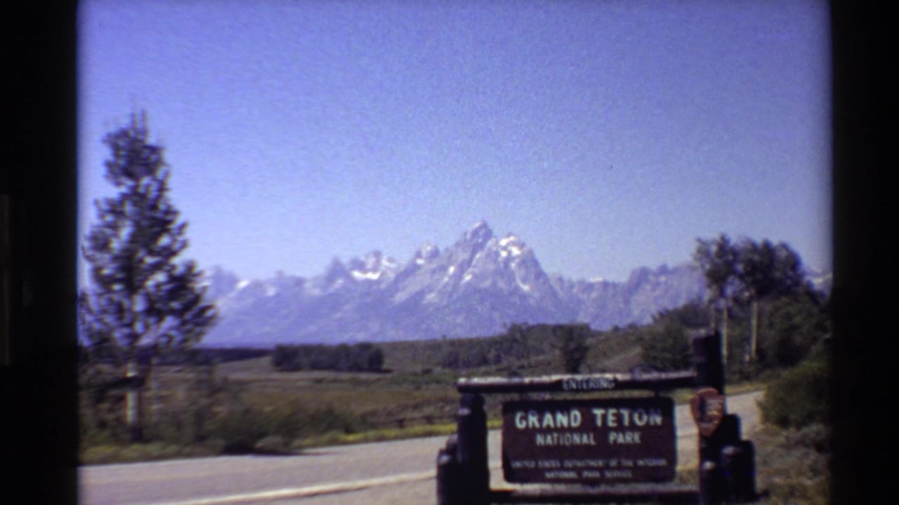 Grand Teton National Park entrance sign beside a road with mountains under a blue sky.