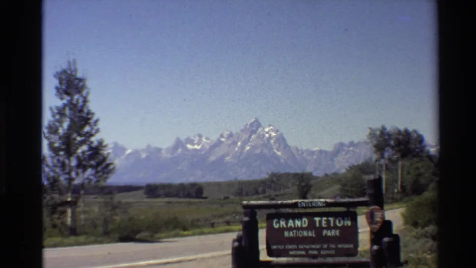 1980 8mm frame centers the Grand Teton National Park entrance sign near a paved road, with a tree at left and the mountain range beyond.