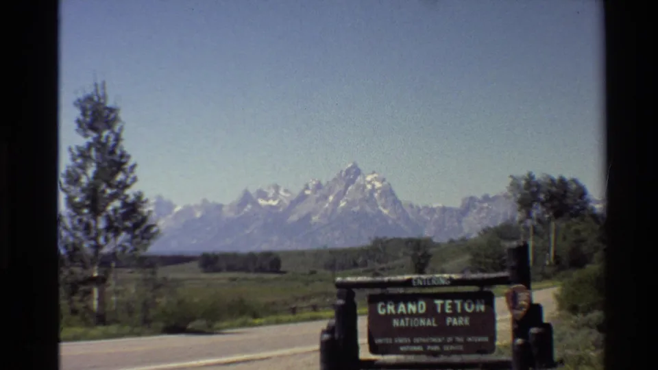 1980 8mm frame holds on the Grand Teton National Park entrance sign, with roadside greenery, a tree, and the Teton range visible beyond.