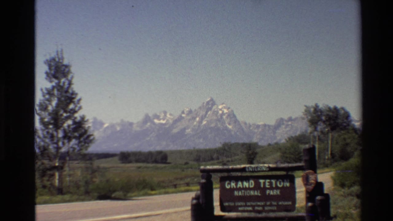 1980s 8mm frame showing the Grand Teton National Park entrance sign, roadside pavement, trees, and snow-capped peaks in the distance.