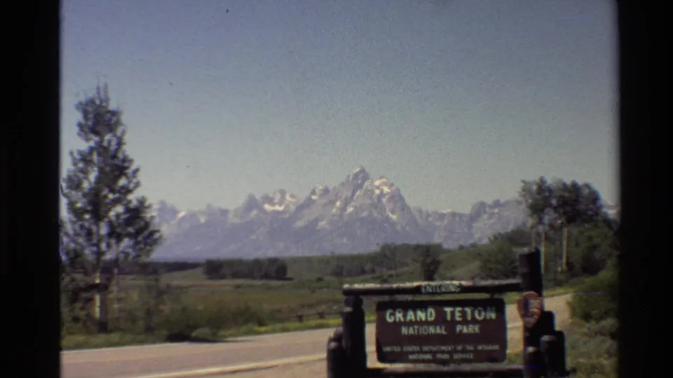 1980s 8mm frame showing the Grand Teton National Park entrance sign, roadside pavement, trees, and snow-capped peaks in the distance.
