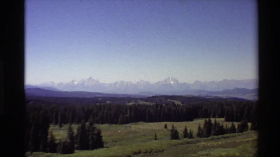 A wide 1980 Western U.S. landscape shows evergreen trees, rolling meadow, distant hills, and snow-capped mountains under pale blue sky.