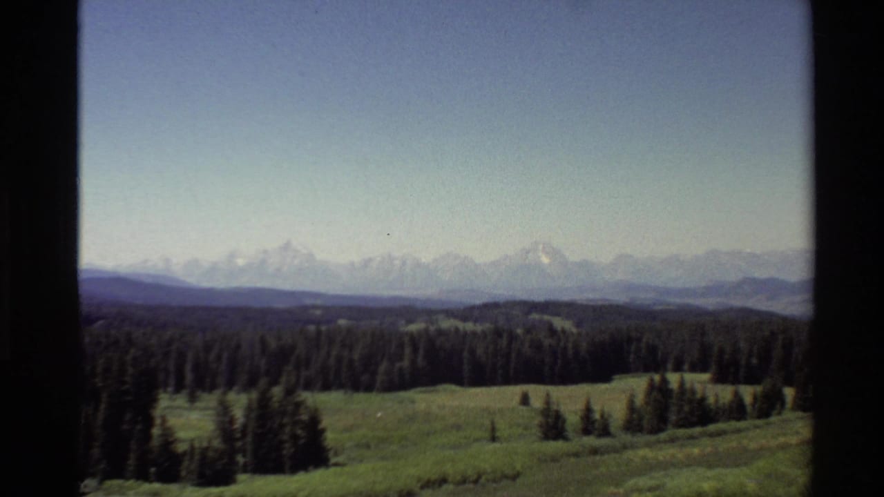 1980 Western U.S. 8mm view of distant snow-capped mountains, pine forest, and meadow framed by a dark window edge.