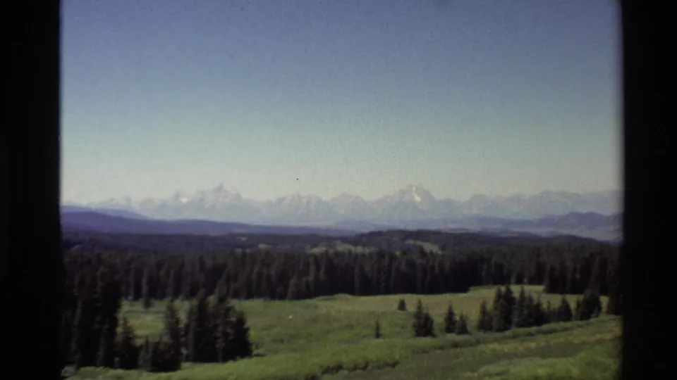 1980 Western U.S. 8mm view of distant snow-capped mountains, pine forest, and meadow framed by a dark window edge.