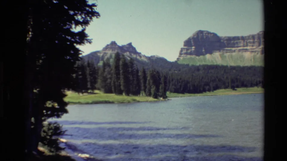 1980 Western U.S. 8mm lakeside view with blue water, pine shoreline, rugged cliffs, and mountain peaks.