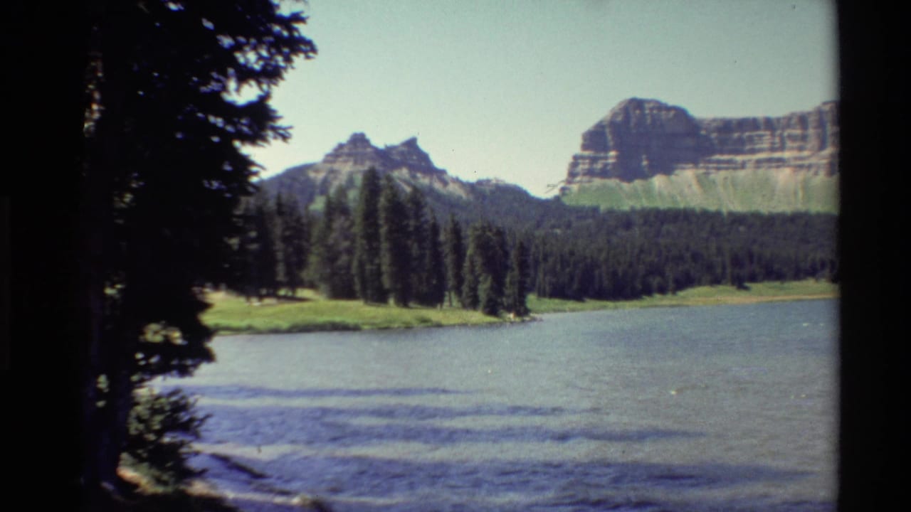 1980 Western U.S. 8mm view across rippled lake water toward evergreen trees and layered rocky cliffs under a pale blue sky.