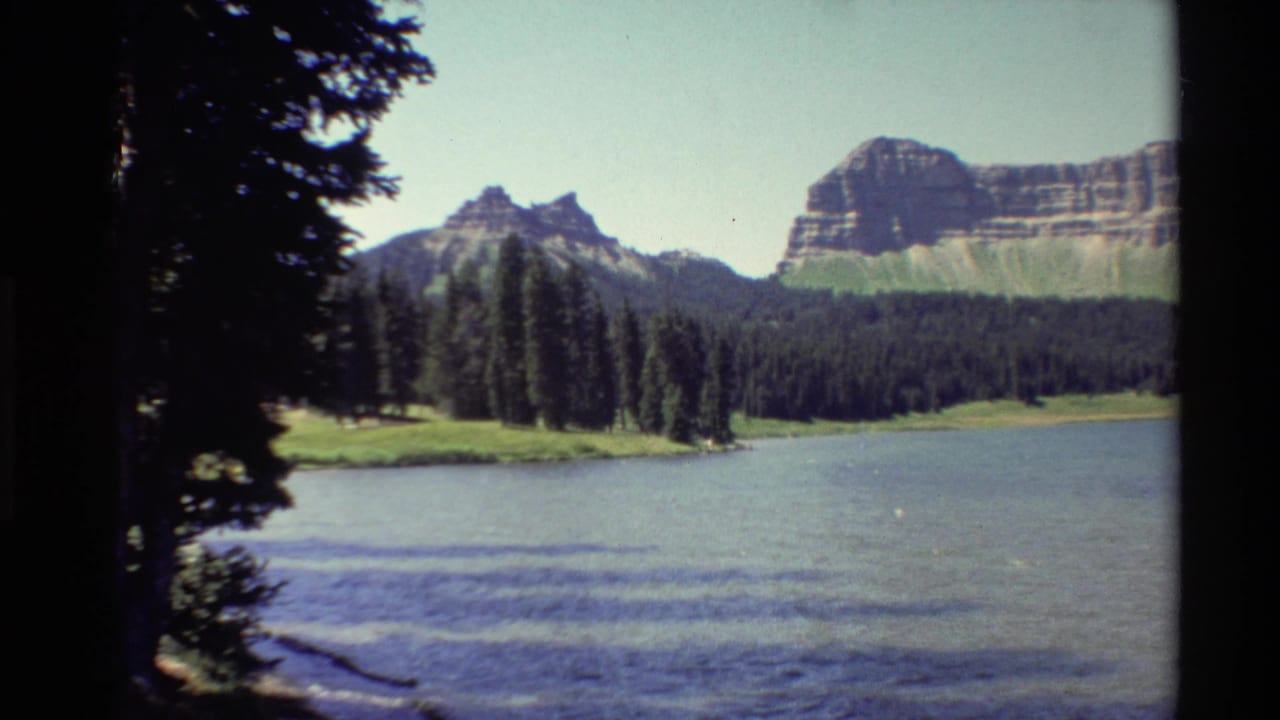 Evergreen trees and rocky cliffs beyond a rippled mountain lake, framed by dark film borders.