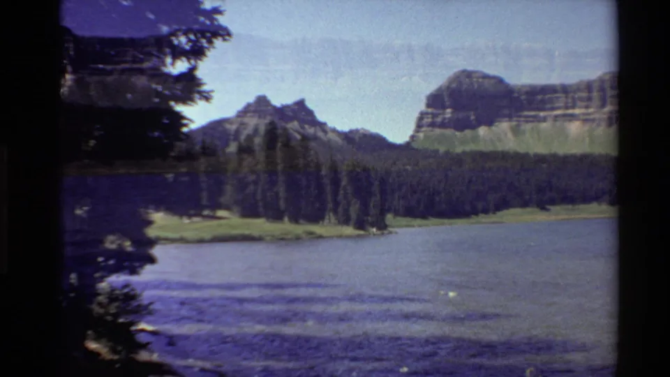 1980 Western U.S. home-movie frame of rippled lake water, a dark evergreen shore, and rocky cliffs, with noticeable 8mm exposure shifts.