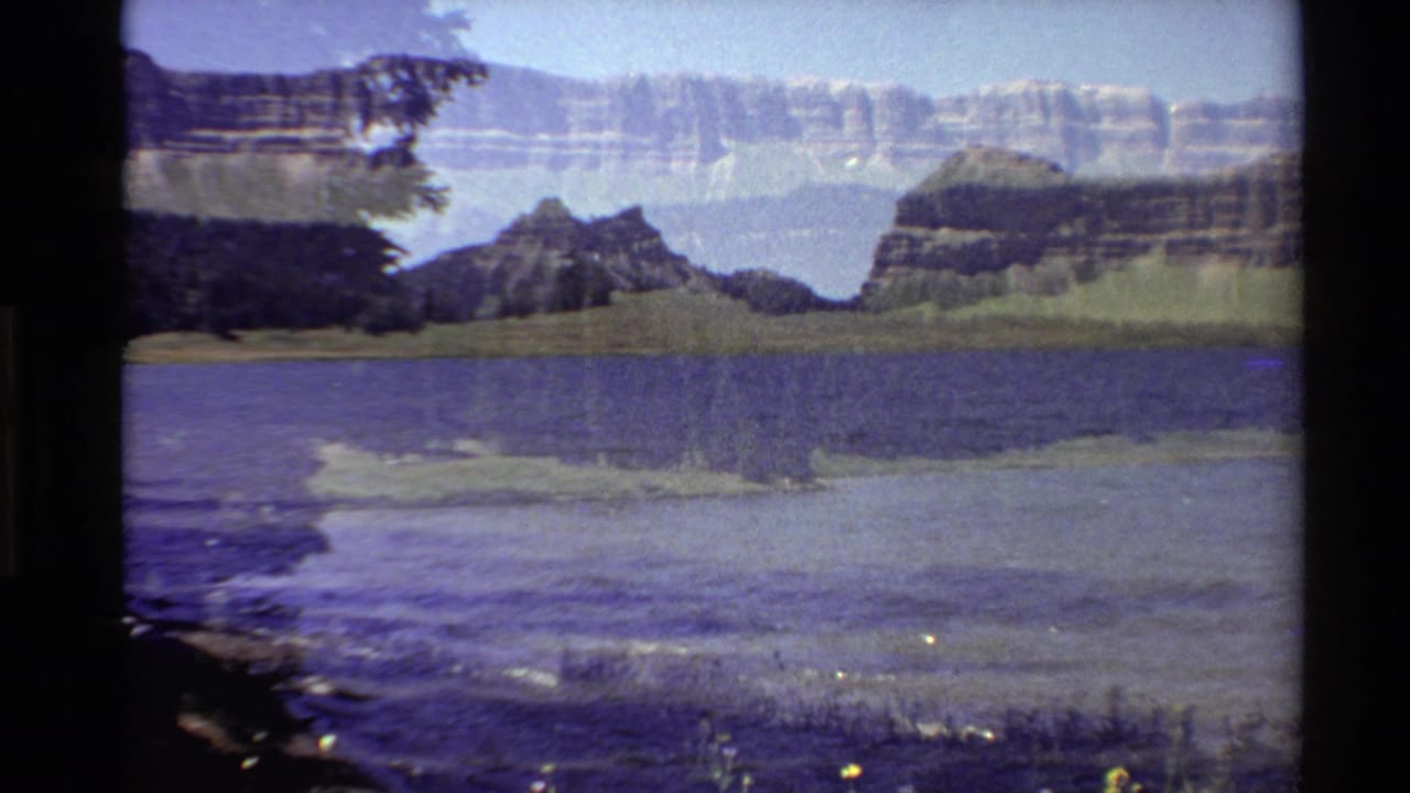 1980 Western U.S. 8mm frame of a lakeshore view with blue water, dark foreground vegetation, forested hills, and distant layered mountains.
