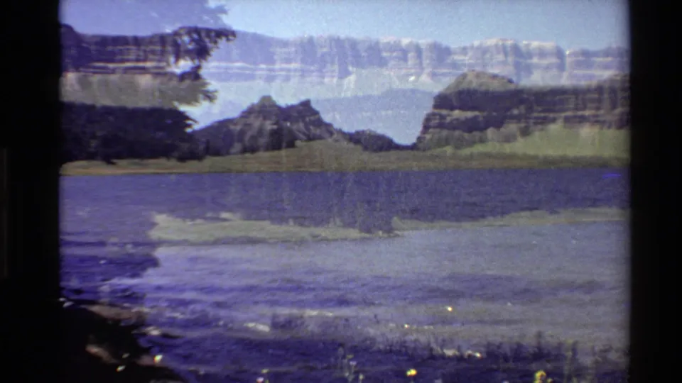 1980 Western U.S. 8mm frame of a lakeshore view with blue water, dark foreground vegetation, forested hills, and distant layered mountains.