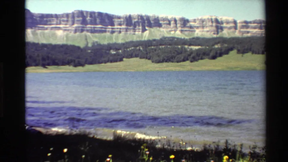1980 Western U.S. home-movie frame showing a broad lake surface with wooded hills and pale, banded mountains under a clear sky.