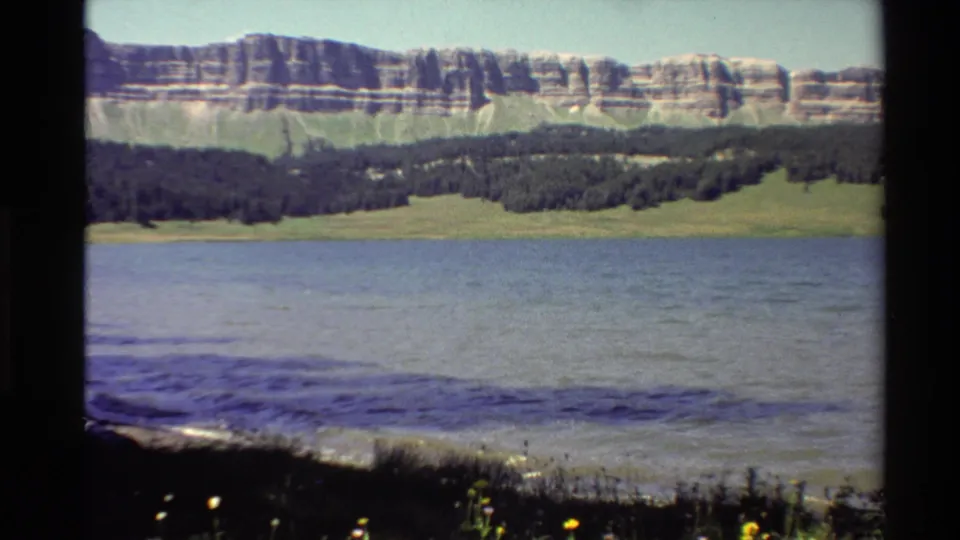 1980 Western U.S. 8mm view across a mountain lake toward green lowlands, dark forest, and a pale banded cliff ridge.