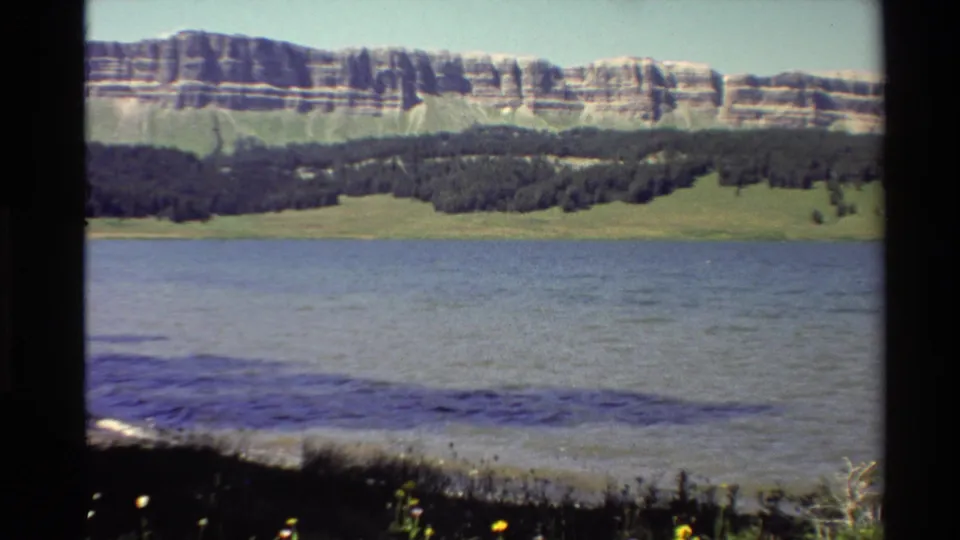 Final 1980 Western U.S. 8mm frame looking over lake water, shoreline plants, rolling forest, and layered mountain cliffs.