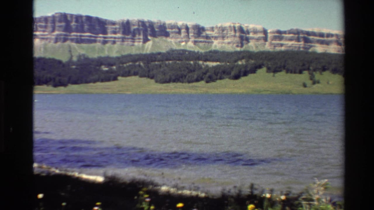 1980 Western U.S. 8mm frame showing rippled water, shoreline plants, a dark tree line, and a layered rocky hillside under clear sky.