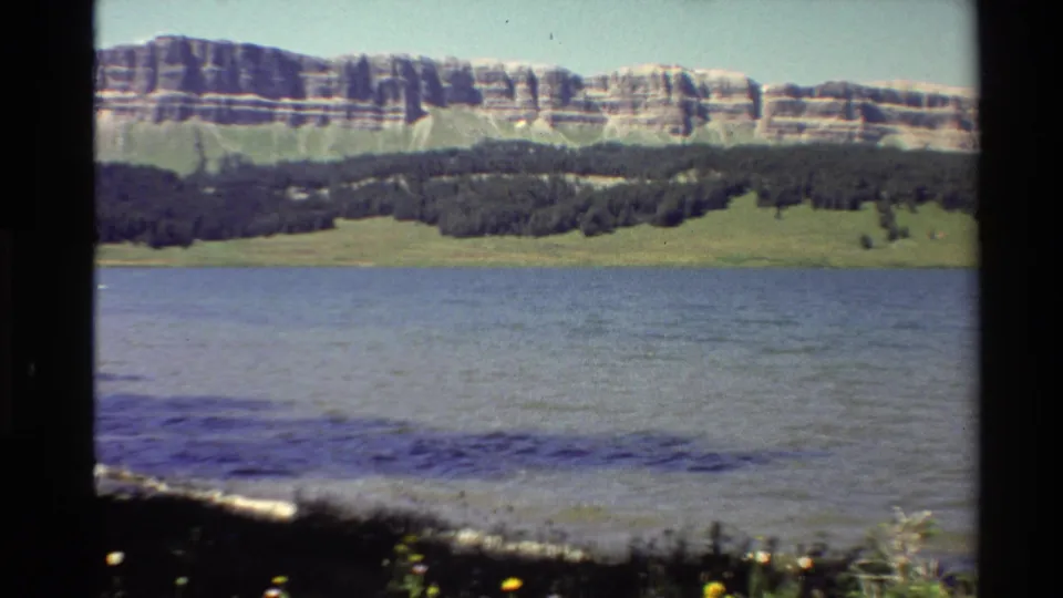 1980 Western U.S. 8mm frame showing rippled water, shoreline plants, a dark tree line, and a layered rocky hillside under clear sky.