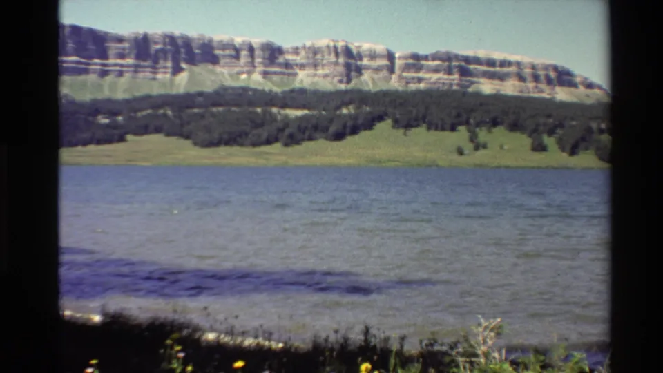 1980 Western U.S. home-movie frame with broad water in front of a green bank, dark trees, and a banded rocky ridge.