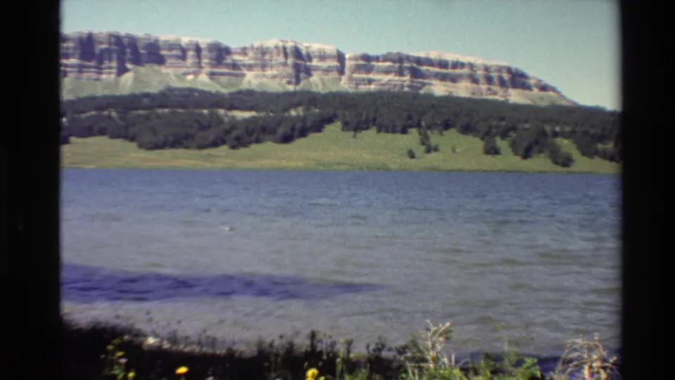1980 Western U.S. 8mm frame holding on rippled water, a low green bank, conifers, and a pale layered cliff in the distance.