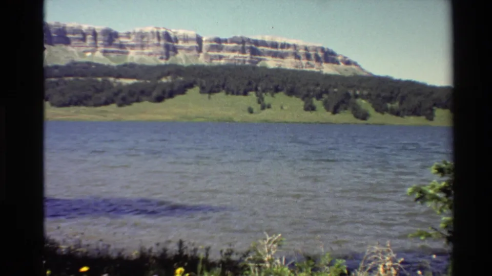 1980 Western U.S. 8mm frame with shoreline vegetation at right, rippled river water, and a rocky hillside beyond trees.