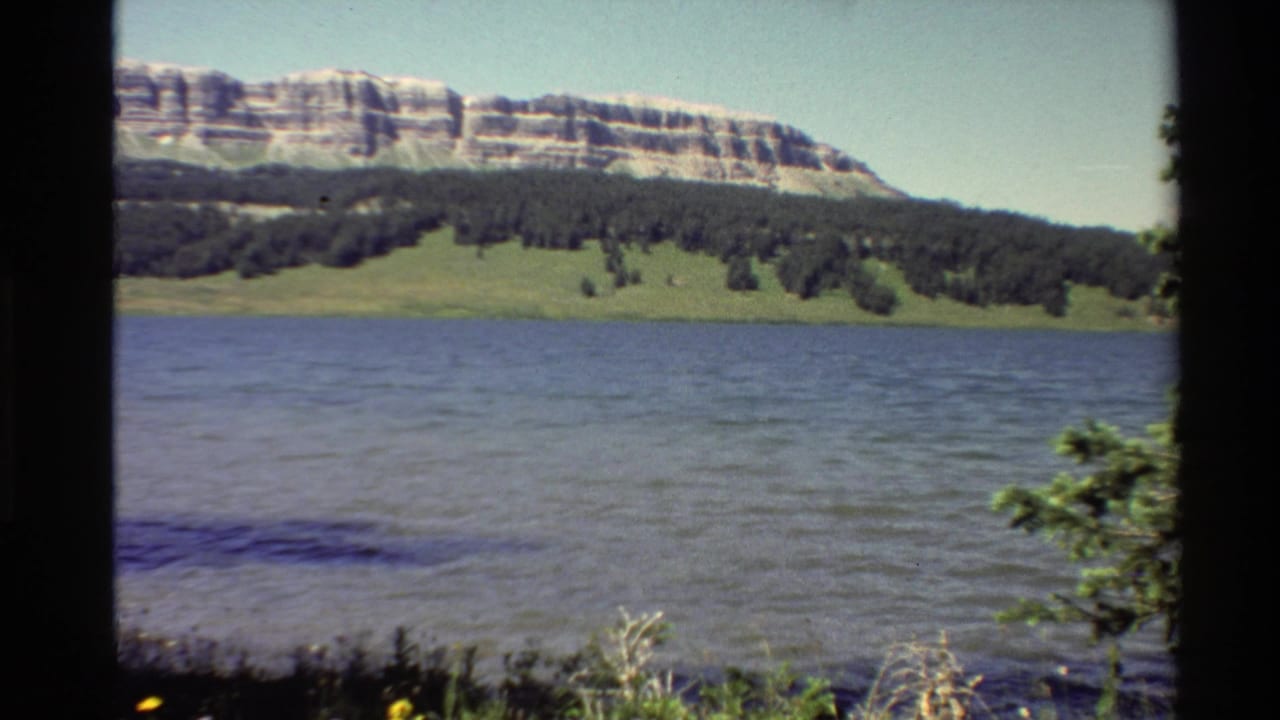 1980 Western USA 8mm frame showing a calm mountain lake, forested shoreline, grassy bank, and layered ridge in bright daylight.