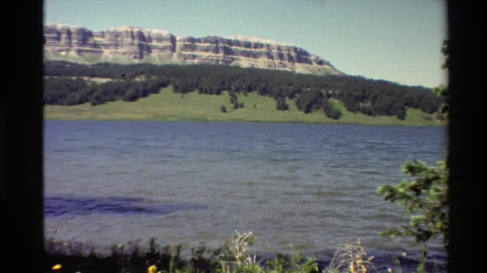 1980 Western USA 8mm frame showing a calm mountain lake, forested shoreline, grassy bank, and layered ridge in bright daylight.