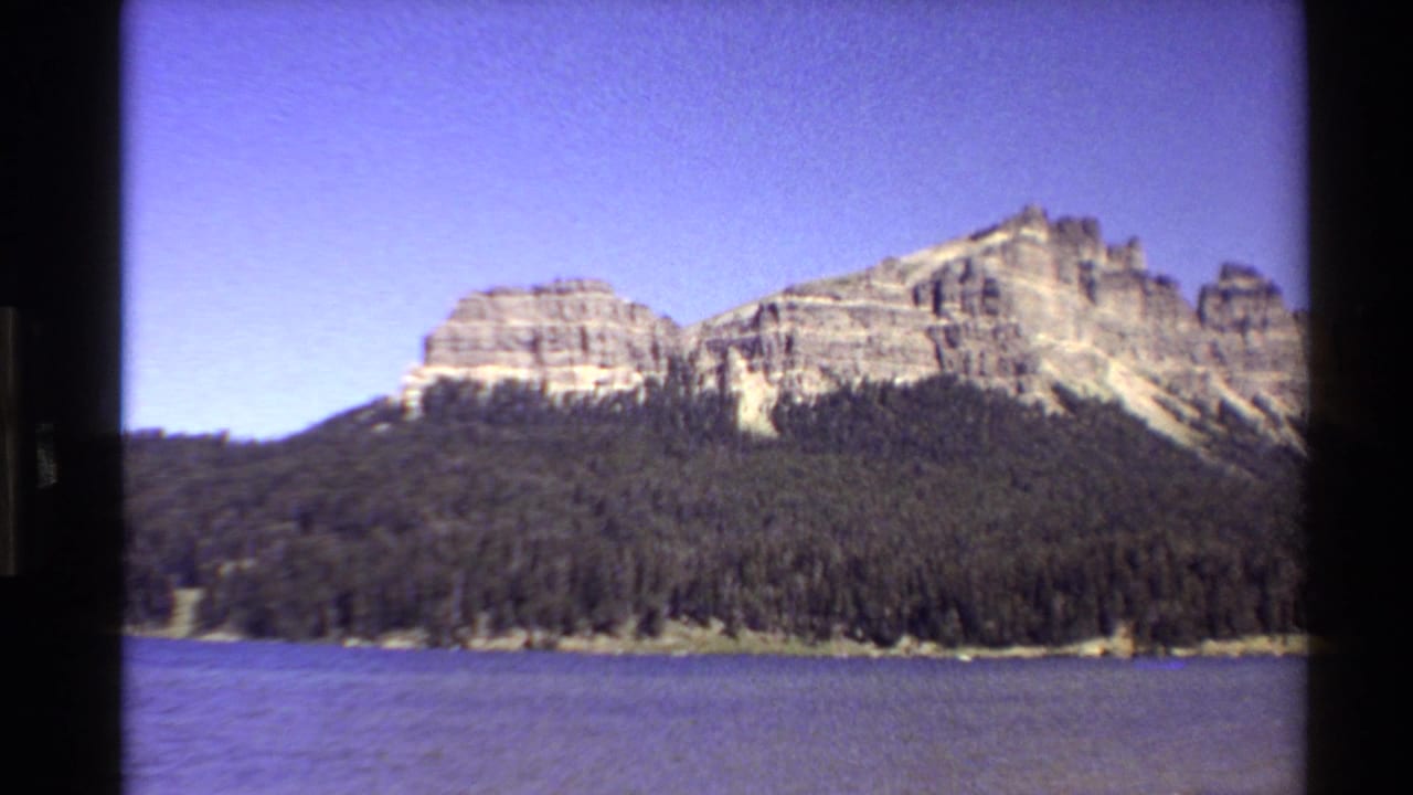 1980 Western U.S. 8mm frame showing a blue lake, evergreen shoreline, and layered rocky cliffs in a steady scenic view.