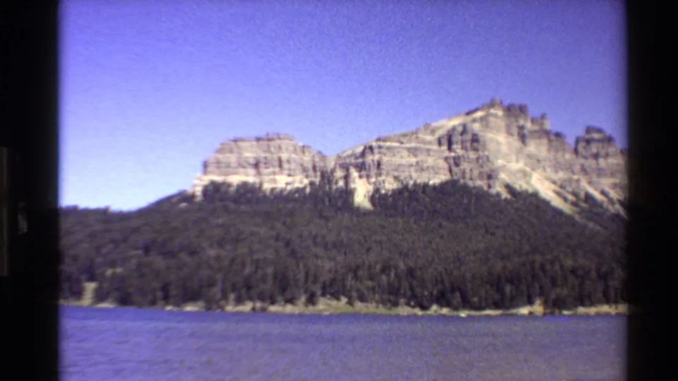 1980 Western U.S. 8mm frame showing a blue lake, evergreen shoreline, and layered rocky cliffs in a steady scenic view.