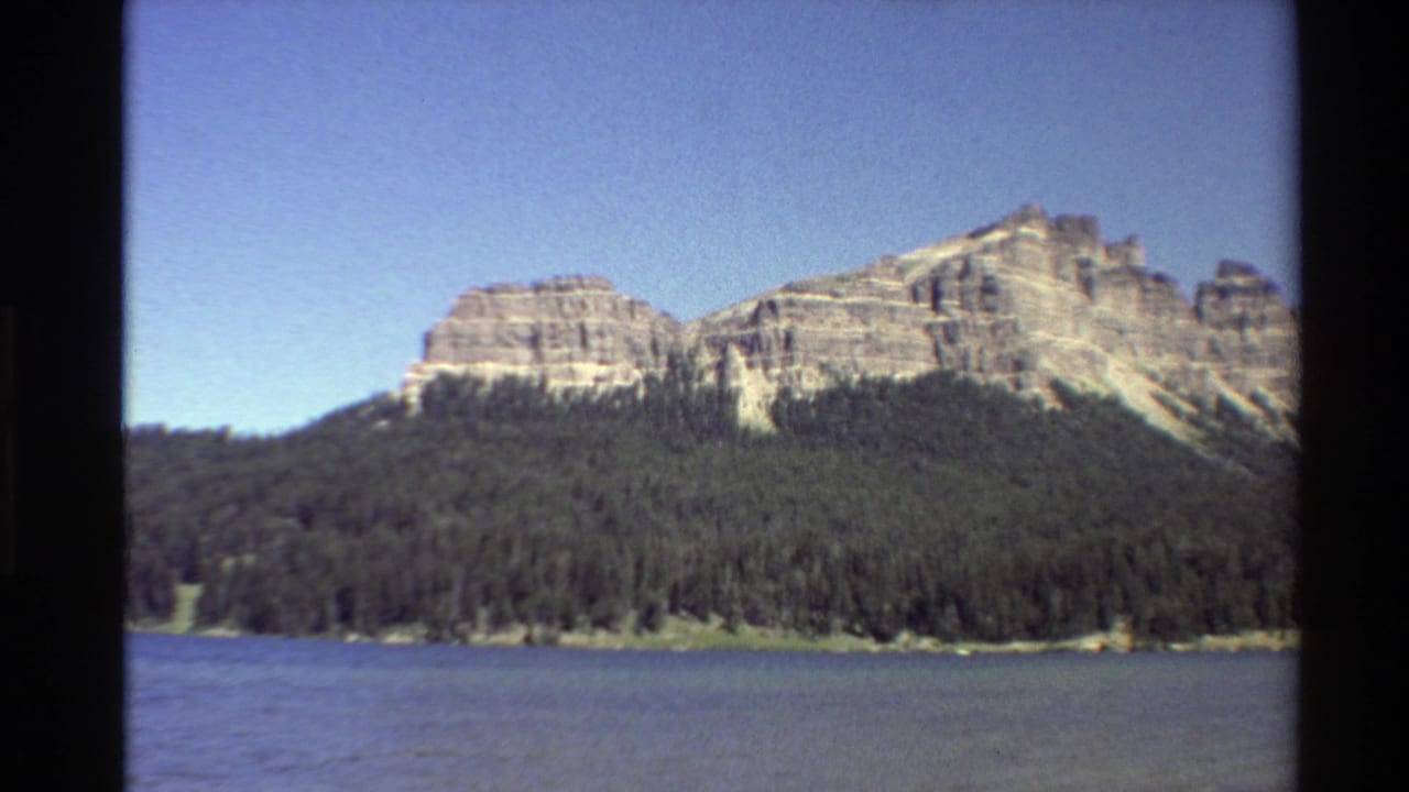 Forested lakeshore with rocky cliffs rising behind it beneath blue sky.