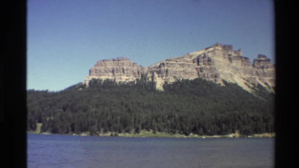 1980 Western U.S. 8mm still with rippled lake water, evergreen trees, and sunlit rock formations in the distance.