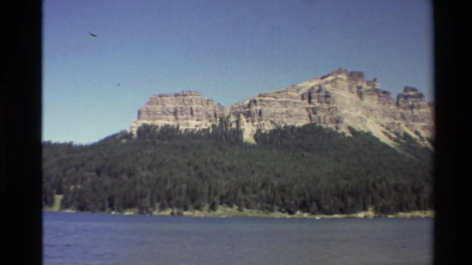 1980 Western U.S. home-movie frame showing lake water, a dense evergreen shore, and layered cliffs with visible film grain.