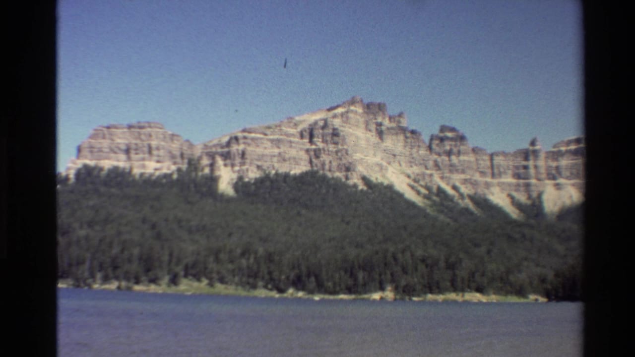 Western U.S. 8mm frame showing a rocky mountain ridge above dense forest and calm blue water in 1980 footage.
