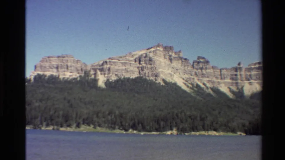 Western U.S. 8mm frame showing a rocky mountain ridge above dense forest and calm blue water in 1980 footage.
