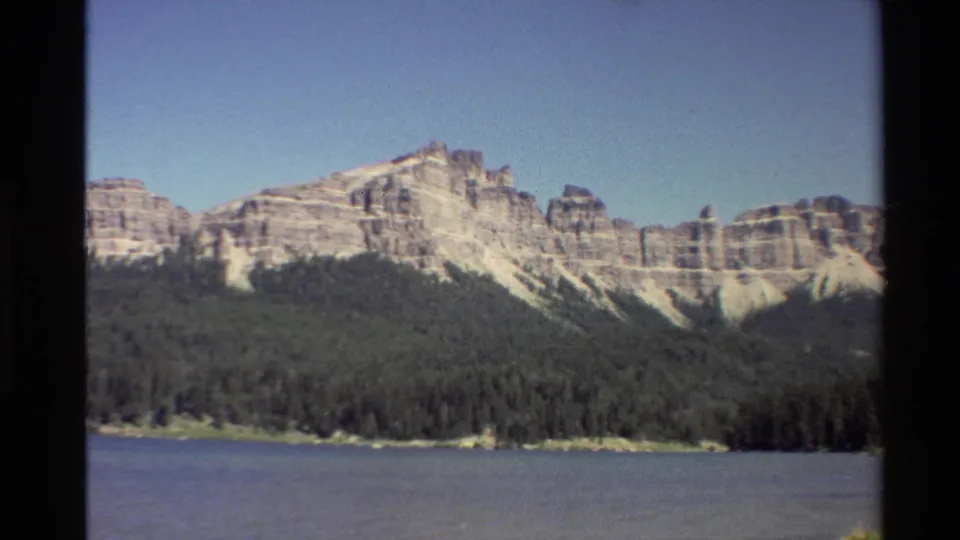 Vintage 8mm view of pale stratified mountain formations over dark evergreen forest and a lake or river.