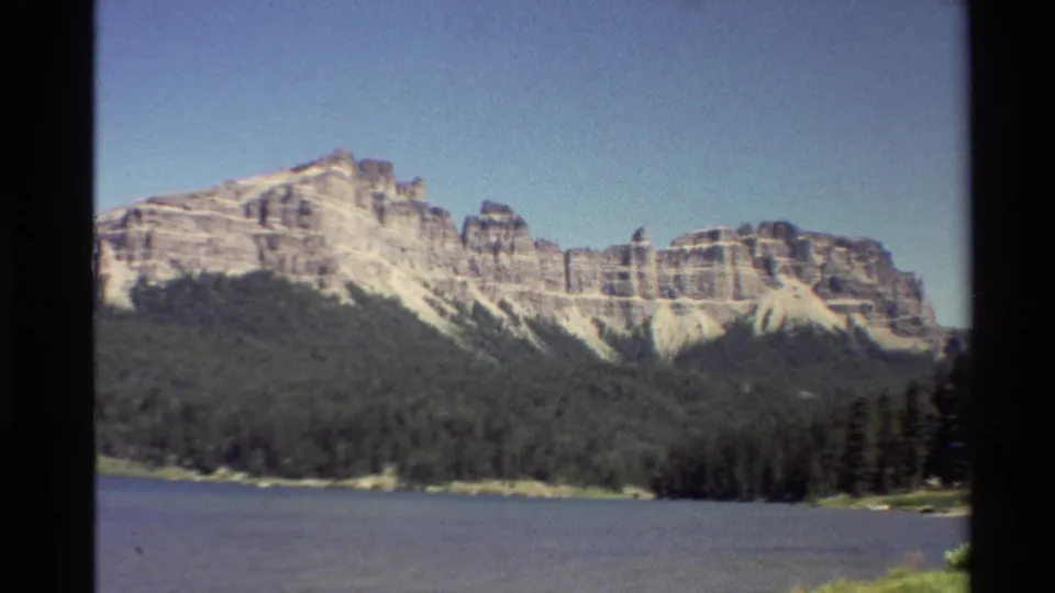 1980 Western U.S. 8mm frame of mountain cliffs, evergreen shoreline, and blue water, framed by dark film edges.