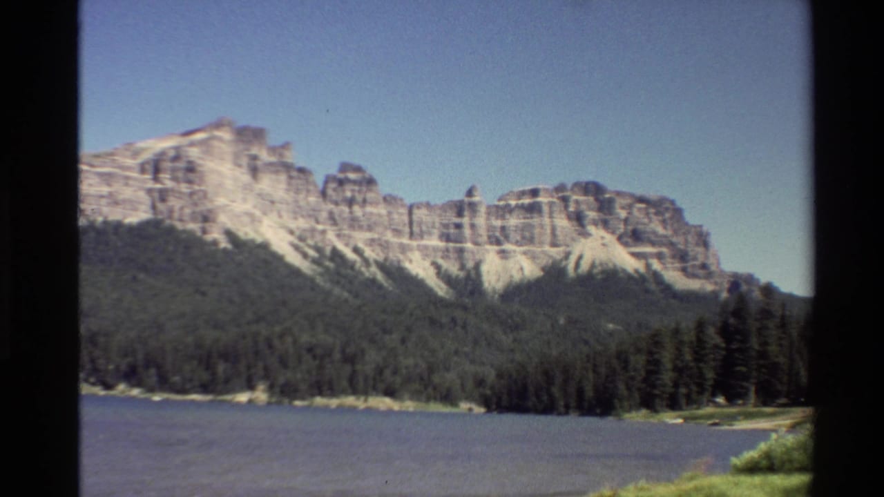 1980 8mm Western USA footage showing pale layered mountains rising behind a calm lake and dark evergreen shoreline.