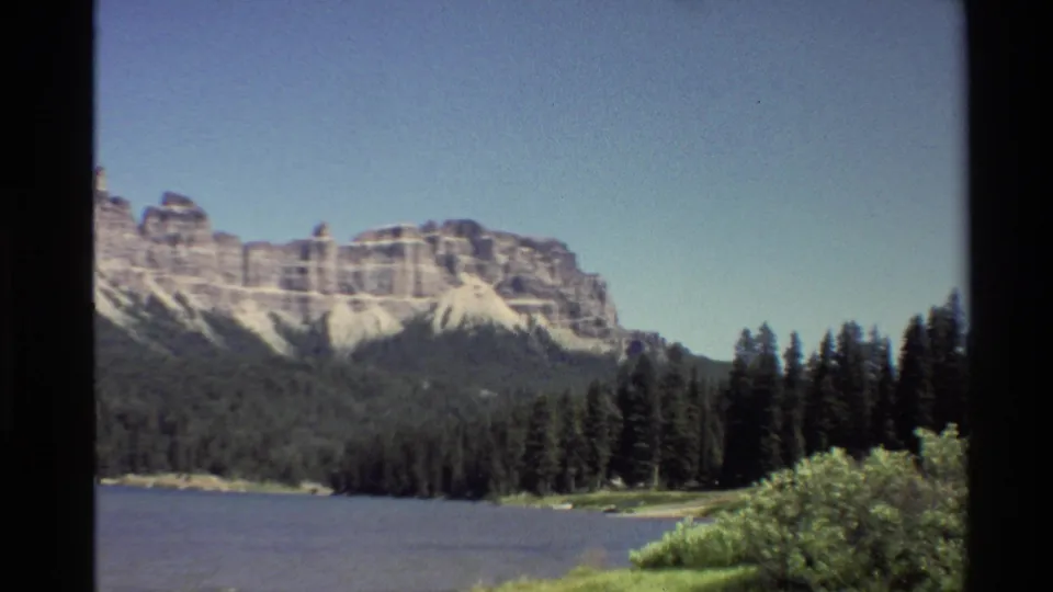 Soft-grained 8mm frame with lake water in front, evergreens along the shore, and a pale rock mountain range beyond.