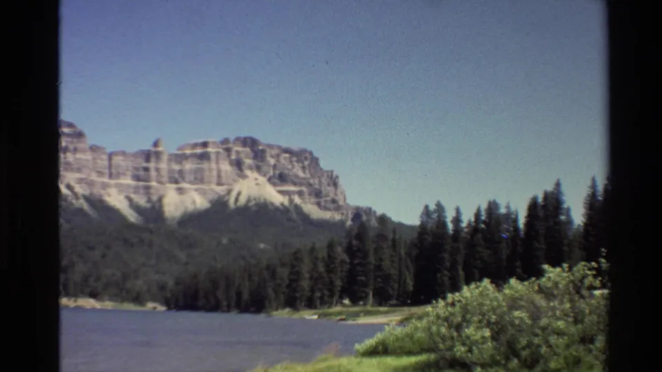 Western USA 8mm landscape frame showing shoreline brush, dark evergreens, lake water, and distant layered mountains.
