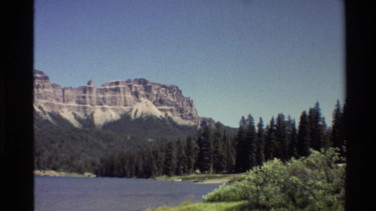 1980 home-movie frame of a Western U.S. mountain range, lake, and pine trees seen through dark 8mm frame edges.
